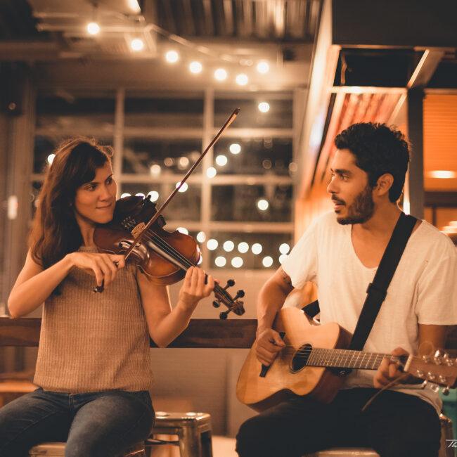 Uma foto de média distância, com iluminação quente e íntima, em um bar ou café à noite, focada em um dueto musical em pé. À esquerda, uma mulher sentada em um banco de metal canta e toca violino, vestindo uma blusa bege texturizada e calça jeans escura. À direita, um homem sentado em um banco similar toca violão acústico e olha para a parceira, vestindo uma camiseta branca de gola V e calça preta com uma alça de violão preta. O fundo é composto por janelas grandes com luzes da cidade embaçadas (bokeh) e uma série de lâmpadas de filamento penduradas em cordas no teto, criando uma atmosfera aconchegante. Entre eles, mais ao fundo, vê-se a silhueta de uma terceira pessoa desfocada, talvez um bartender ou cliente. A câmera está no nível dos olhos e a profundidade de campo é rasa, mantendo os músicos em foco nítido.