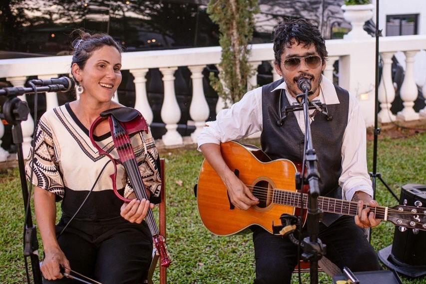 Fotografia em plano médio de um duo musical se apresentando ao ar livre. À esquerda, a violinista Gabriela Lançarin canta de olhos fechados em um microfone de pedestal enquanto segura um violino elétrico vermelho estruturado. Ela veste uma blusa com estampas geométricas em tons de preto e bege. À direita, um homem de óculos escuros, camisa social branca e colete preto toca violão acústico e canta em seu próprio microfone, além de usar uma gaita de boca presa em um suporte no pescoço. Ao fundo, vê-se a fachada de um prédio residencial de dois andares em tom amarelado, árvores e uma caixa de som em primeiro plano à esquerda.