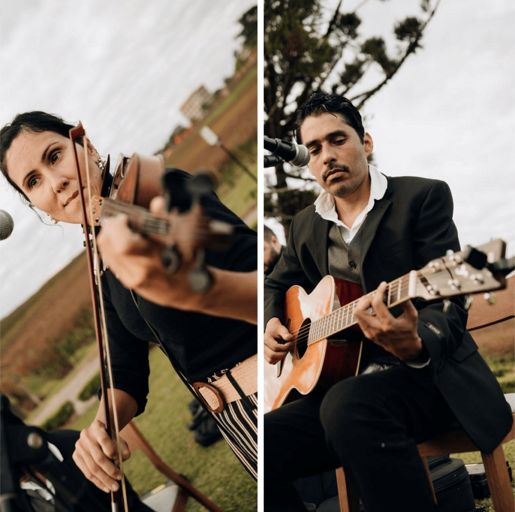 Apresentação musical do TNG Duo em casamento ou cerimonial ao ar livre. À esquerda, close-up de uma violinista concentrada segurando o arco. À direita, um músico de terno escuro toca violão acústico. Ao fundo, paisagem verde desfocada sob céu nublado.